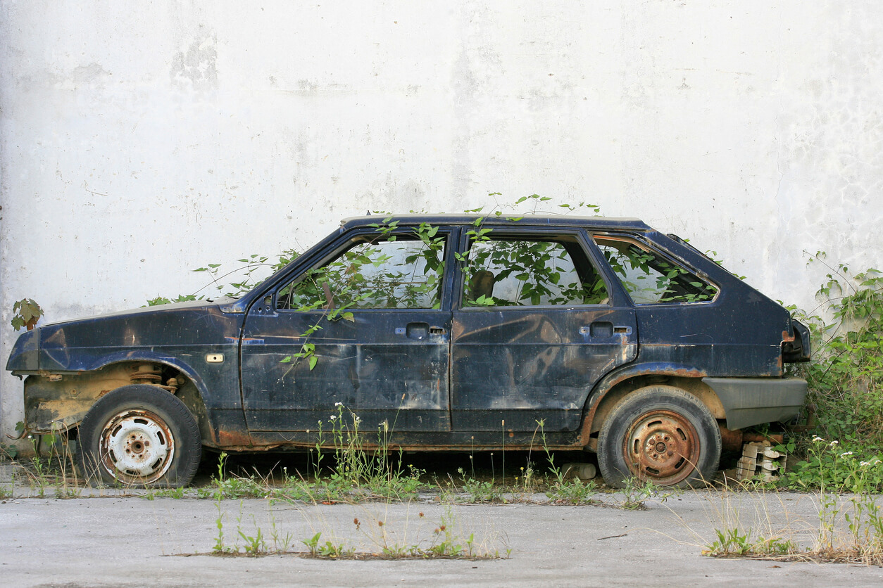 Discarded old car in overgrown grass next to the wall.